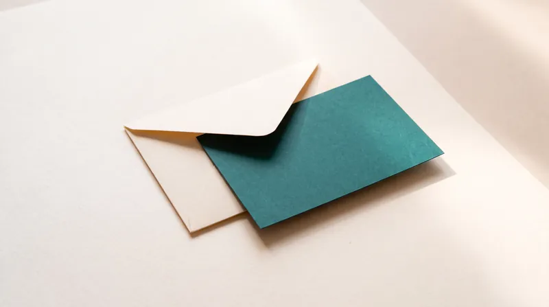 A cream paper envelope with its flap open and a blank teal card sliding out.